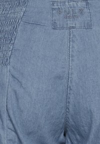 Primer plano de una tela de mezclilla azul con un parche cosido que dice "PULZ JEANS" y un detalle elástico gathered en el lado.