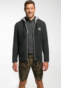 Gilet tricoté en charbon avec capuche grise, fermeture éclair et poches latérales. Associé à un lederhosen marron avec embossage floral et boutons.