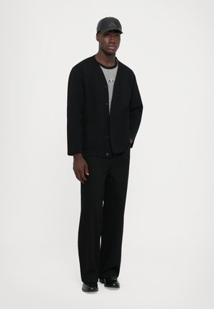 Cardigan noir avec des boutons, associé à une chemise grise et un pantalon noir à jambes larges. Le modèle porte également une casquette noire et des chaussures. Design minimaliste.