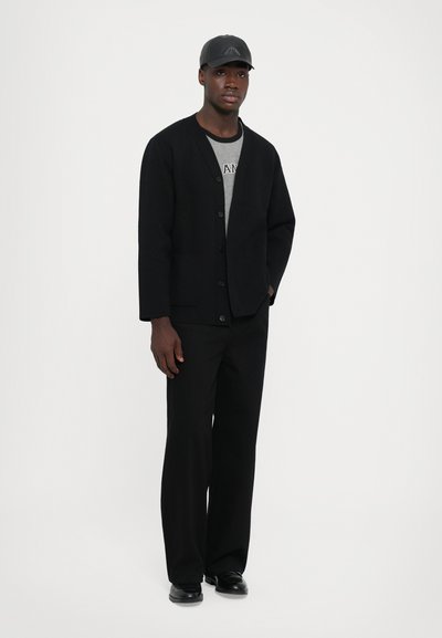 Musthave muströstsest mustvalge kampsun nööpidega, mis on kombinatsioon halli särgi ja laia mustade pükstega. Mudel kannab ka musta mütsi ja kingi. Minimalistlik disain.