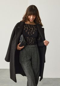 Czarny płaszcz na czarnej koronkowej bluzce, w zestawie z szarymi, strukturalnymi spodniami. Trzyma czarną kopertówkę. Miękkie, gładkie faktury z nowoczesnym wyglądem.