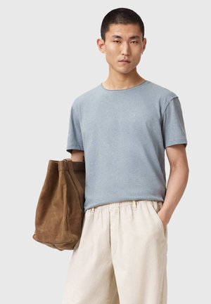 Nuori mies, jolla on vaaleansininen T-paita ja beiget housut, pitää suurta ruskeaa kantolaukkua kädessään ja seisoo yksinkertaisen vaalean taustan edessä.