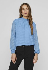 Blusa azzurra chiara con collo alto arricciato, maniche lunghe e texture a strisce a zigzag. Abbinata a pantaloni neri, ha una vestibilità ampia.