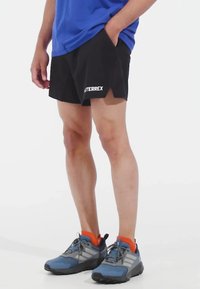 Black athletic shorts with a loose fit, side pockets, and white "TERREX" branding on the lower left. Paired with blue and gray athletic shoes.
