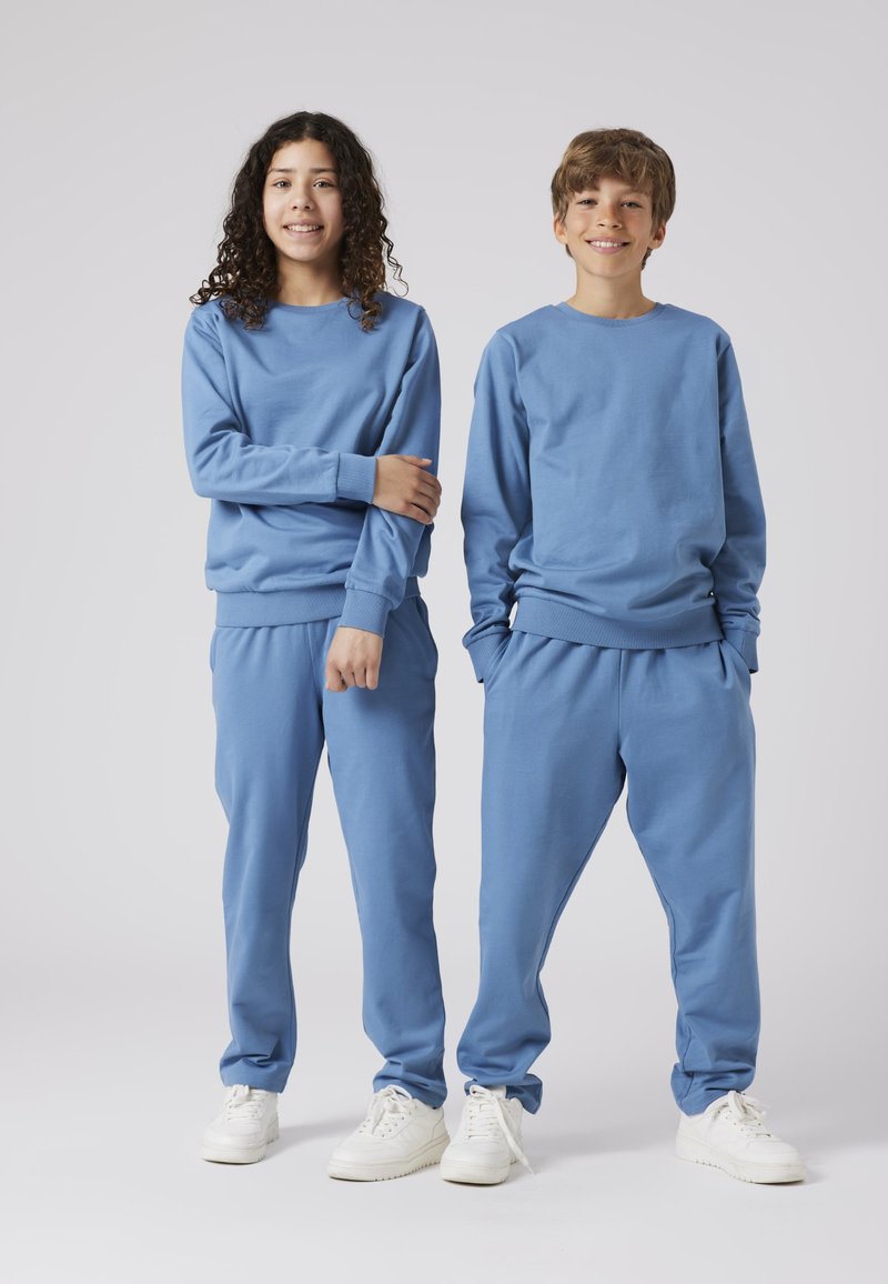 Gaiši zils treniņu krekls un bikses komplekts, kas izgatavots no mīkstas kokvilnas, ar ribotām aprocēm un jostu, papildināts ar baltām sporta apaviem.