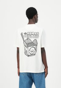 Hvitt bomull t-skjorte med et grafisk motiv av en takeaway nudleboks med spisepinner og teksten "ADIDAS ORIGINALS" på baksiden. Løs passform.