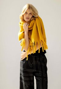 Gelber Wollschal mit fransigen Kanten, um die Schultern gelegt, erg�änzt durch schwarze Hosen mit weißen geometrischen Linienmustern.