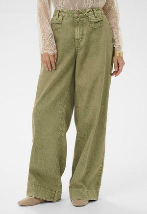 Nő széles szárú olívazöld nadrágban, zsebes fedőrésszel, csipkés hosszú ujjú felsőben, bézs hálós cipőben és arany karkötőben.