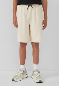 Beige Baumwollshorts mit einem elastischen Bund, schwarzem Kordelzug und Seitentaschen, die knapp über dem Knie enden, kombiniert mit grauen Turnschuhen.