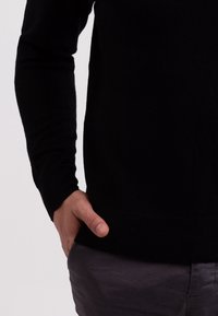 Schwarzer, gestrickter Pullover mit langen Ärmeln und ribbeligem Saum. Hand in der Taille platziert, kombiniert mit grauen Hosen. Glatte Textur, einfaches Design.