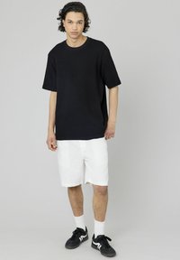 Pull noir à manches courtes côtelé associé à un short blanc. Des baskets noires et des chaussettes blanches complètent la tenue. Fond uni.