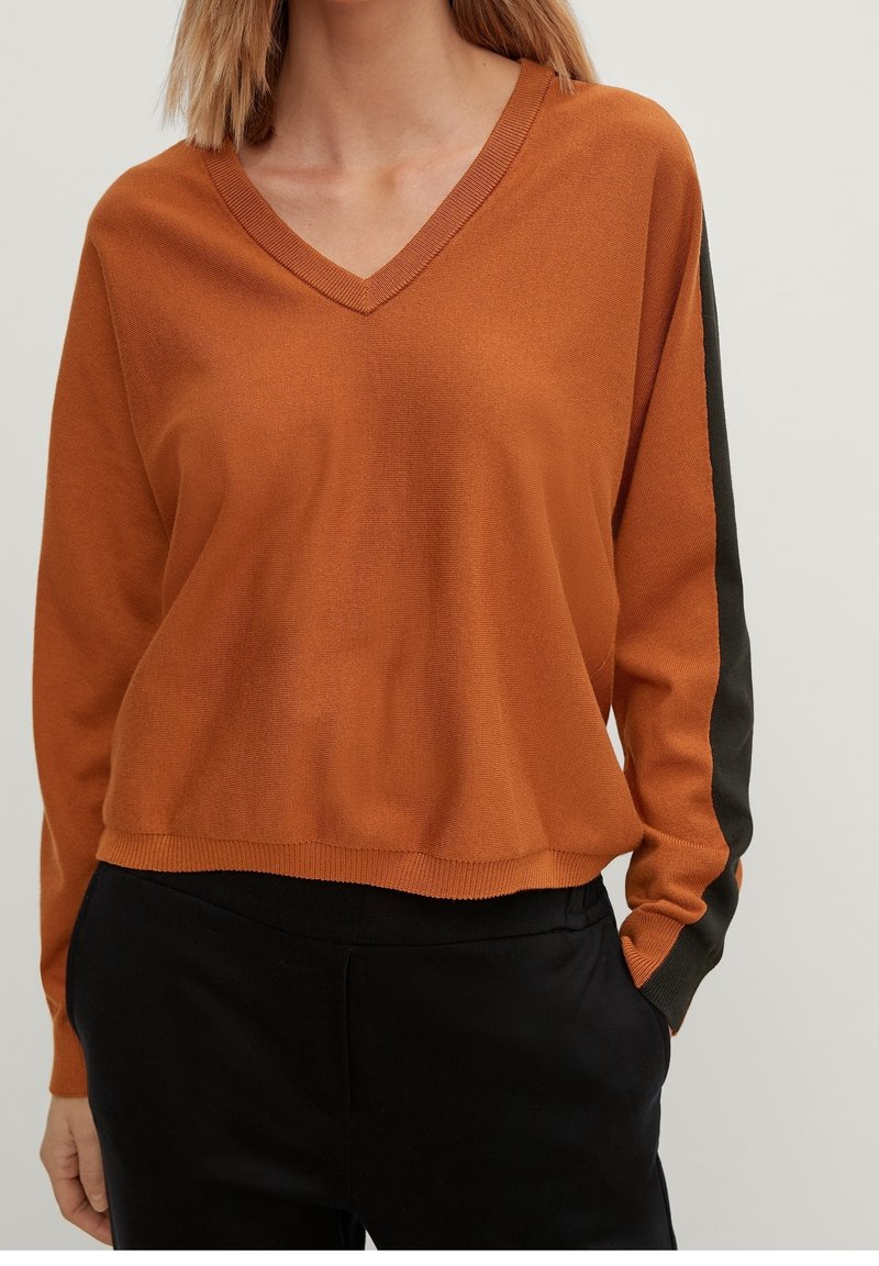 Suéter marrón de cuello en V con mangas oscuras contrastantes, textura de punto, corte relajado y bajo con puños, combinado con pantalones negros.