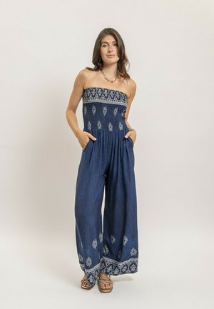 Femme portant une combinaison bleu marine sans bretelles avec des motifs floraux blancs et des sandales dorées, debout les mains dans les poches sur un fond neutre.