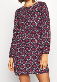 Donna che indossa un abito a maniche lunghe sopra il ginocchio con uno sfondo blu navy e un motivo floreale rosso, in piedi con le mani rilassate ai lati.