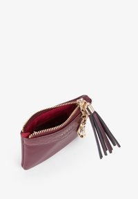 Portafoglio in pelle bordeaux con accessori dorati, chiusura a zip e dettaglio con nappina nera. L'interno presenta una fodera in tessuto rosso.