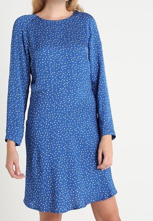 Femme portant une robe bleue à manches longues, longueur genou, avec des pois blancs irréguliers, se tenant devant un fond clair uni.