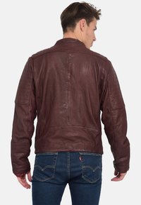 Dunkelburgunderfarbene Lederjacke mit glatter Textur, ausgestattet mit gesteppten Schulterakzenten und einem klassischen Kragen. Getragen über blauen Jeans.