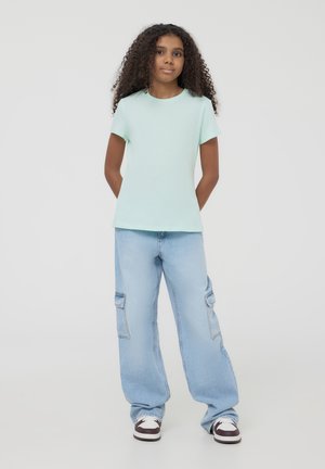 T-shirt mint verde a maniche corte abbinato a pantaloni cargo larghi azzurri. I pantaloni presentano tasche laterali e una vestibilità rilassata, indossati con sneakers.