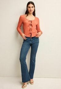 Oranges strukturiertes Langarmoberteil mit einer Bindeschleife vorne, kombiniert mit dunkelblauen Schlagjeans und hellen Schuhen.