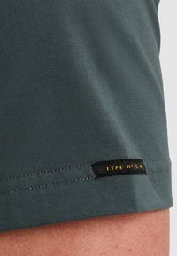 Grå stof T-shirt med en glat tekstur, der har et sort mærke i kanten med gul tekst til identifikation.