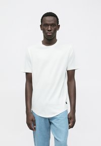 Bílé tričko s krátkým rukávem a kulatým výstřihem, doplněné světle modrými džínami. Tričko má zaoblený lem a minimalistický design.