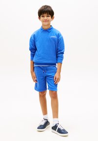 Αγόρι όρθιο, φοράει μπλε φούτερ με μακριά μανίκια και σορτς που ταιριάζουν, με αθλητικά παπούτσια, σε λευκό φόντο.