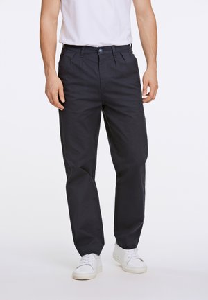 Man draagt zwarte geplooide broek, witte casual sneakers en een wit T-shirt dat in de broek is gestopt, staande op een effen lichte achtergrond.