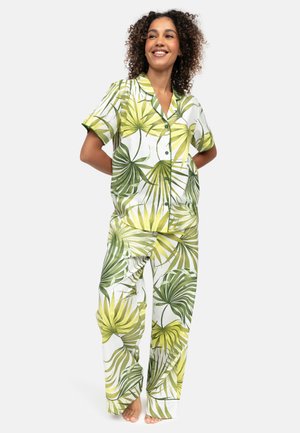 Lachende vrouw met krullend haar, gekleed in een overhemd met korte mouwen en een tropische bladprint in groen en geel, en bijpassende wijduitlopende broek.