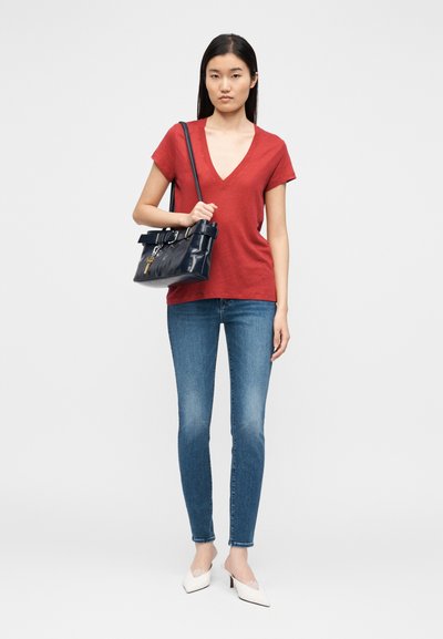 T-shirt vermelha com decote em V, jeans skinny azuis, saltos brancos sem tiras e uma mala navy com detalhe de cinto, segurada na mão direita.