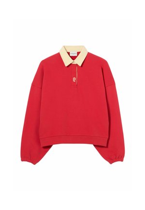 Giovane donna in piedi con una mano in tasca, indossa un maglione rosso oversize, una camicia gialla con colletto, jeans blu, cintura marrone e stivali neri.