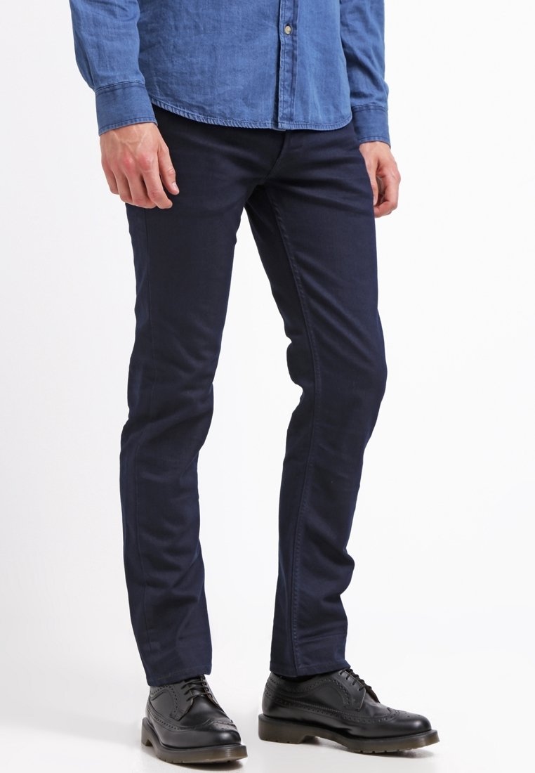 Tummansininen kapealahkeinen housu, jonka pinta on sileä, viisi taskua ja tavalliset vyölenkit. Yhdistettynä mustiin nahkakenkiin.