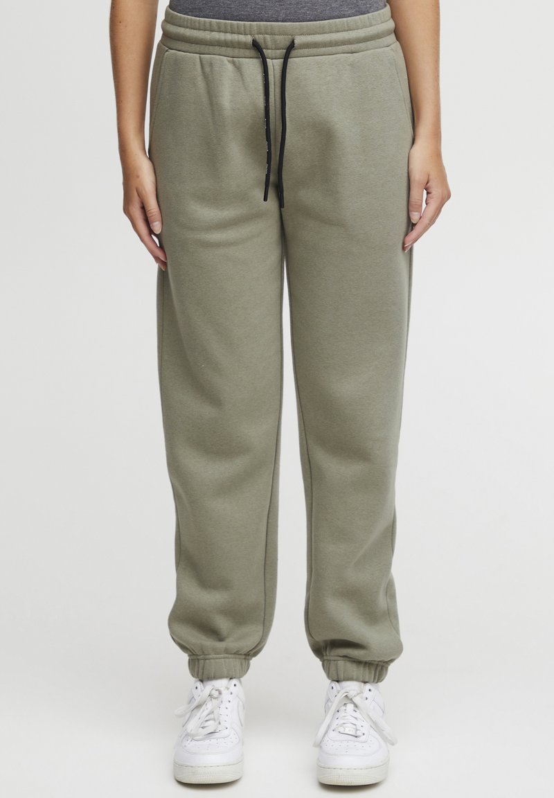Olivgrüne Sweatpants mit elastischen Bündchen, verstellbarem schwarzen Kordelzug und Seitentaschen, kombiniert mit weißen Sneakers. Weiches Baumwollmischmaterial.