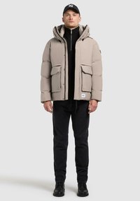 Beige puffer jacket with hood, two front pockets, and a zipper. Worn over a black high-neck top and paired with dark jeans and black boots.