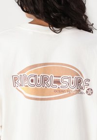 Camiseta oversized de algodón blanca con un diseño brillante en marrón y dorado de una tabla de surf que presenta el texto "RIP CURL SURF OCEAN TECHNOLOGY".