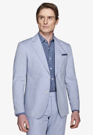 Mężczyzna ubrany w jasnoniebieski garnitur z dopasowaną marynarką i spodniami na ciemnoniebieskiej koszuli z guzikami, patrzący lekko w bok.