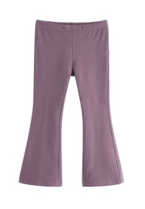 Ung pige iført en lilla hue, lyserød bluse med lange ærmer, lilla bukser og mørkerøde sneakers, stående med hænderne i lommerne mod en hvid baggrund.