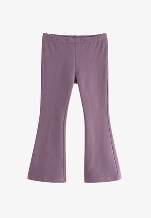 Ung pige iført en lilla hue, lyserød bluse med lange ærmer, lilla bukser og mørkerøde sneakers, stående med hænderne i lommerne mod en hvid baggrund.
