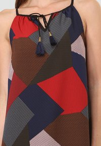 Top sin mangas multicolor con patrones geométricos en rojo, azul marino, marrón y blanco. Tiene un escote en V con un lazo y dos borlas azul marino.