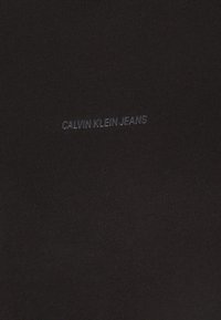 Schwarz geripptes Material mit einem subtilen "CALVIN KLEIN JEANS" Logo in einer etwas dunkleren Nuance, platziert im unteren Bereich.