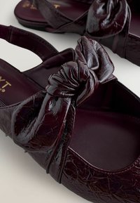 Burgundy lackläder slingback-lägenheter med knuten frontdetalj och texturerad finish som visas på en ljus yta.