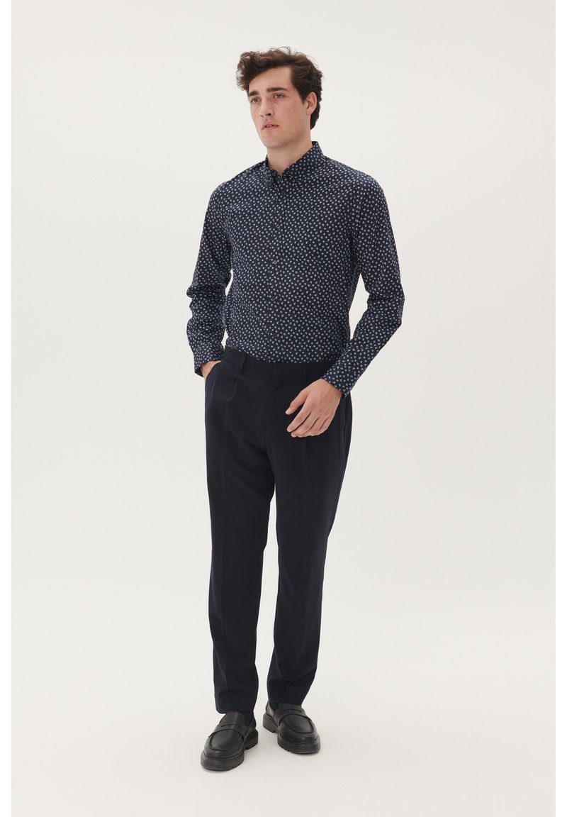 Marineblauwe overhemd met een wit patroon, gecombineerd met donkere nette broeken. Het shirt heeft een kraag en lange mouwen. Zwarte schoenen maken het geheel af.