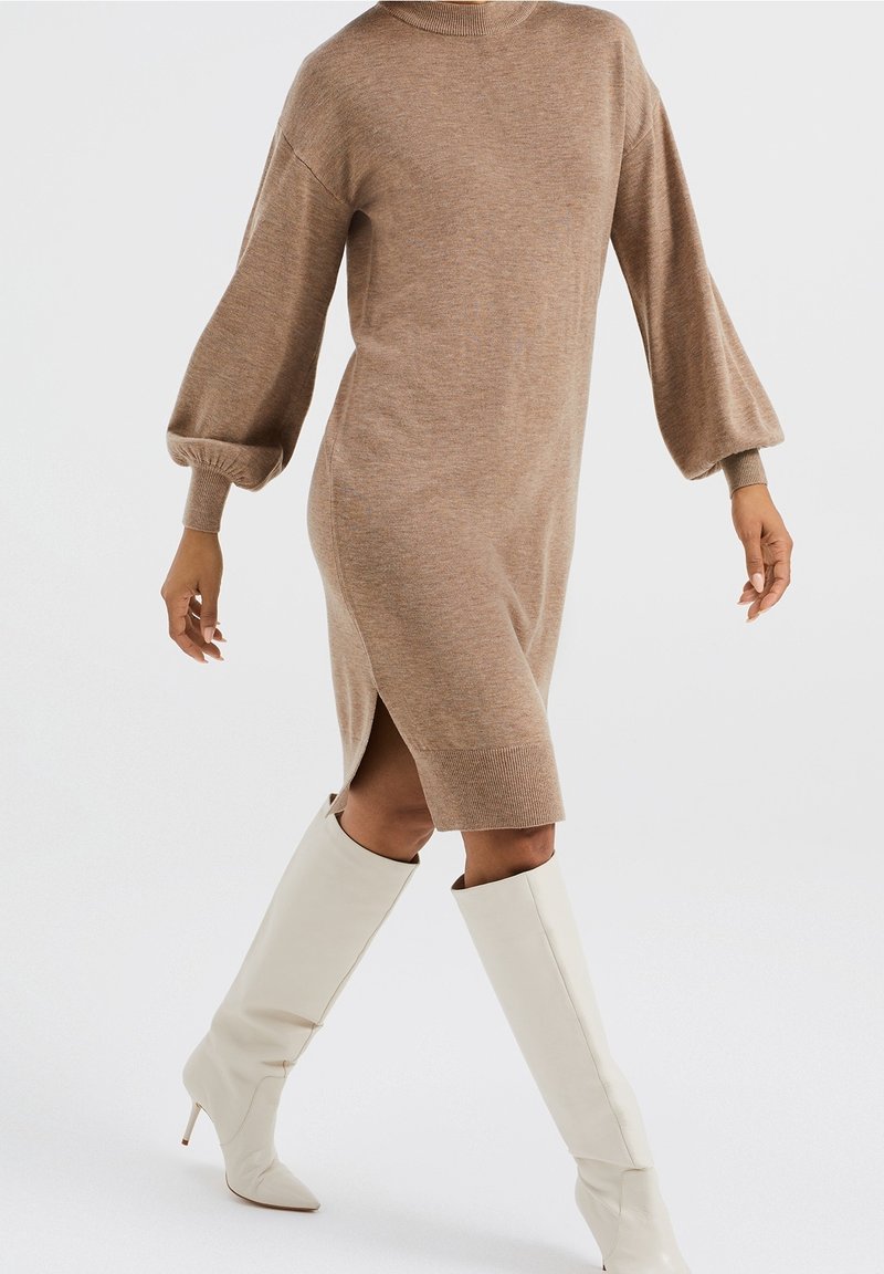 Robe en maille à manches longues marron avec fente latérale portée avec des bottes blanches à talons montant jusqu'aux genoux, sur une personne en marche sur fond uni.