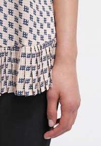 Ljusmönstrad topp med en krämfärgad bas och marinblå geometriska former, en plisserad fåll och mjuk tygtextur. Svarta byxor synliga.