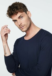 Marineblaues Langarmhemd aus leichtem Stoff, mit Rundhalsausschnitt und engen Bündchen. Haut sichtbar am Unterarm mit Tätowierung.