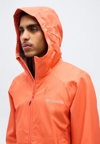 Άνδρας που φοράει πορτοκαλί αδιάβροχο μπουφάν με κουκούλα της Columbia, κοιτάζοντας προς τα αριστερά, με την κουκούλα σηκωμένη και το φερμουάρ μερικώς κλειστό, μπροστά από απλό φόντο.