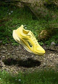 Zapatilla deportiva amarilla con parte superior de malla, punta redondeada y un swoosh negro. Cuenta con una entresuela texturizada y suela de goma para mayor agarre.