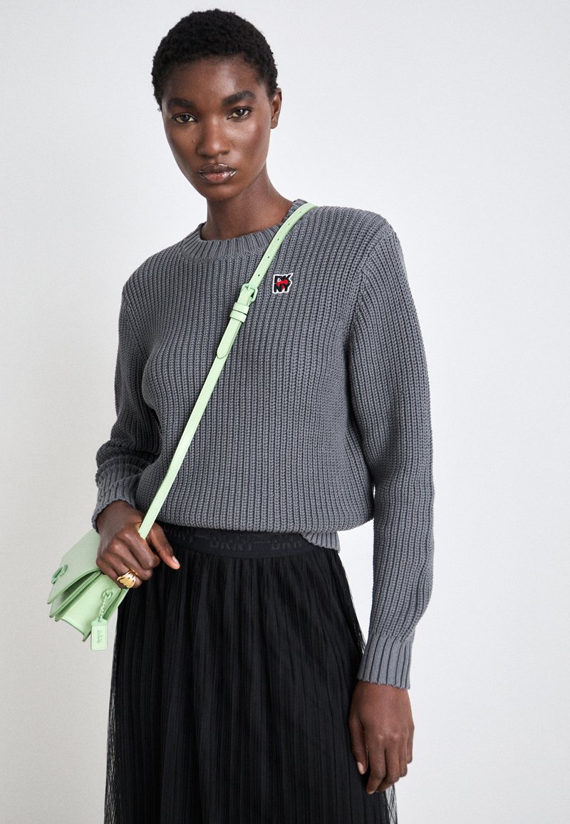 Grau gestreifter Pullover mit Rundhalsausschnitt und Logo-Patch, kombiniert mit einem schwarzen Faltenrock. Eine hellgrüne Umhängetasche setzt einen Farbklecks.