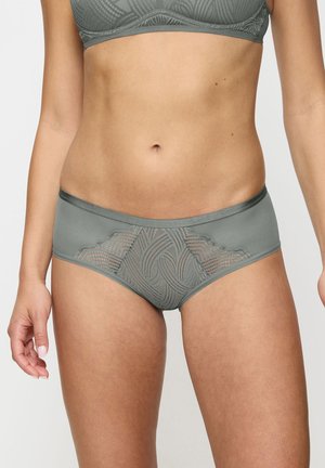 Femme portant des sous-vêtements gris à motif de dentelle et un soutien-gorge assorti, visible du milieu du torse jusqu'aux cuisses sur un fond uni.