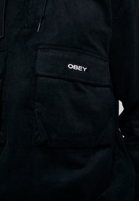 Black corduroy jacket with a front pocket featuring an "OBEY" logo. The material has a ribbed texture and a modern, practical design.