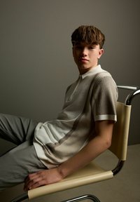 Kortærmet polo-shirt med et tekstureret mønster i creme og taupe, med en knapkrave. Model siddende i en stol i metal og læder.